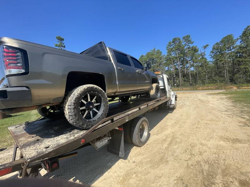 Gray pickup loaded on a flatbed on a sandy dirt lot with pine trees behind