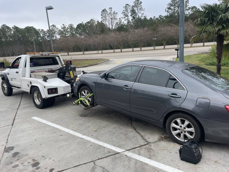 White wrecker with a gray sedan raised on its wheel lift in a parking lot